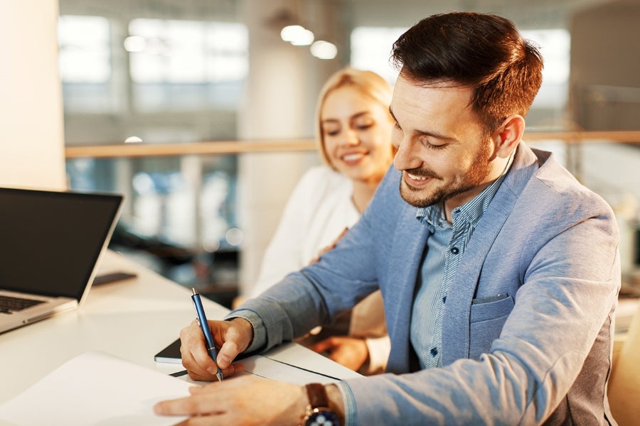 Smiling man signing documents with woman beside him.